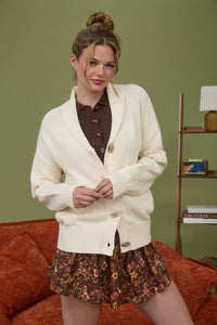 Woman wearing a white cardigan over a brown shirt and floral skirt in a room with a green wall and red couch.