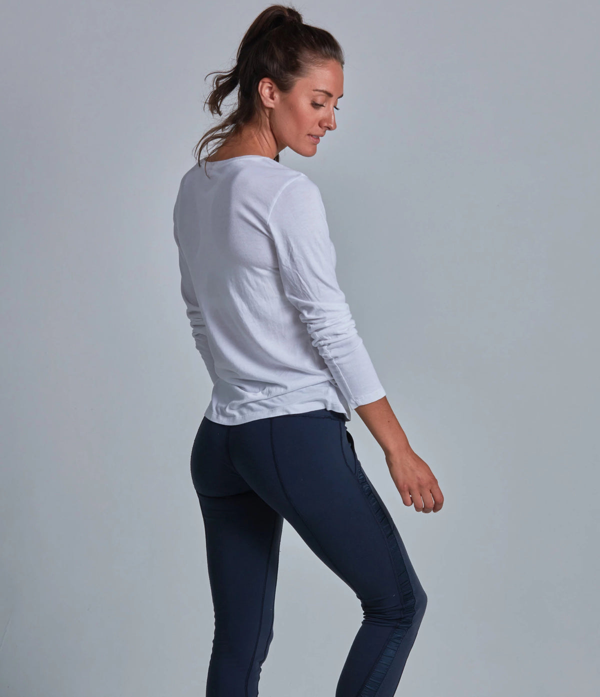 Woman wearing a white long-sleeve top and dark blue leggings against a gray background