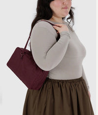 Woman wearing a gray long-sleeve top and brown skirt, holding a maroon handbag against a white background