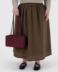 Person wearing a long brown skirt holding a maroon handbag on a white background
